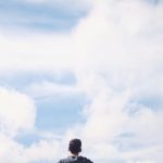 From below back view of anonymous male traveler standing with reached arms enjoying cloudy sky during vacation