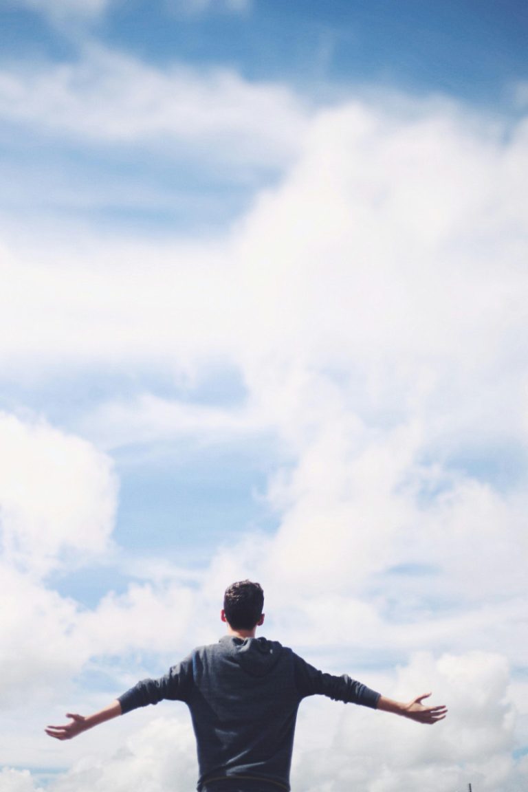 From below back view of anonymous male traveler standing with reached arms enjoying cloudy sky during vacation
